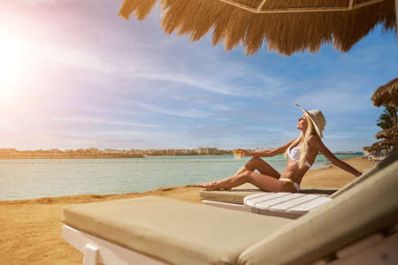 Sexy woman wearing bikini sitting on lounger under straw canopy umbrella at the beach holding glass...