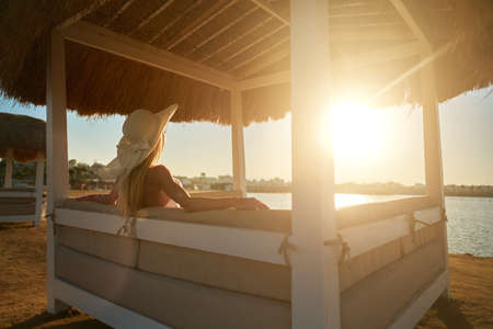 Woman sitting at Cabana with straw roof on a sandy beach on sunsetの写真素材