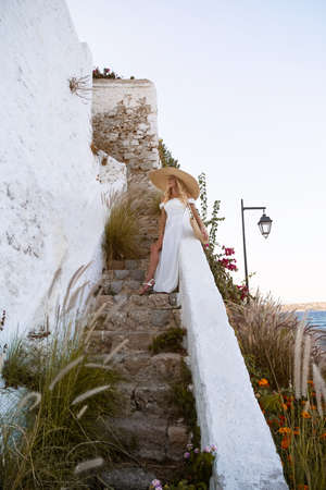 young elegant woman wearing straw hat and white dress standing on stars at old town - Greece,...