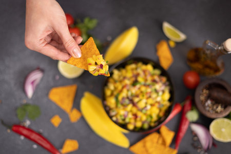 Freshly made mango salsa in a black ceramic bowl, nacho chips and ingredientsの写真素材