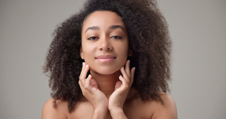 Beauty and healthcare concept - beautiful African American woman with curly afro hairstyle and clean, healthy skin touching face with hand, doing facial massage, posing and looking at cameraの写真素材