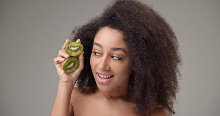 Beauty, vitamins and healthcare concept - beautiful African American woman with curly afro hairstyle and clean, healthy skin holding kiwi halves in her hands posing and looking at the cameraの写真素材