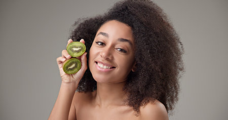 Beauty, vitamins and healthcare concept - beautiful African American woman with curly afro hairstyle and clean, healthy skin holding kiwi halves in her hands posing and looking at the cameraの写真素材