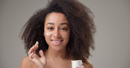Beauty and healthcare concept - beautiful African American woman with curly afro hairstyle and clean, healthy skin applying moisturizer to her face with a slight smile and positive moodの写真素材