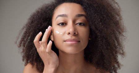 Beauty and healthcare concept - beautiful African American woman with curly afro hairstyle and clean, healthy skin applying moisturizer to her face with a slight smile and positive moodの写真素材