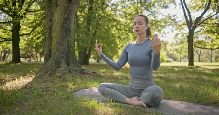 A young beautiful white girl practices yoga in the park and meditates in the lotus position among the trees in a clearing on a summer dayの写真素材