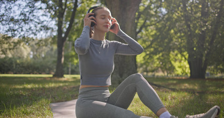 A young beautiful white girl practices yoga in the park and meditates in the lotus position among the trees in a clearing on a summer day and listens to music from her phone with headphonesの写真素材