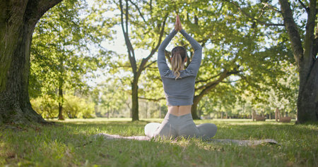 A young beautiful girl practices yoga in the park and meditates in the lotus position among the trees in a clearing on a summer dayの写真素材