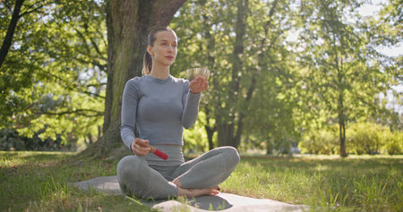 A young beautiful girl doing yoga in the park and meditating in the lotus position among the trees in a clearing on a summer day and using a singing brass bowlの写真素材