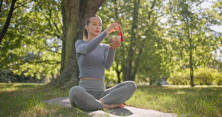 A young beautiful girl doing yoga in the park and meditating in the lotus position among the trees in a clearing on a summer day and using a singing brass bowlの写真素材