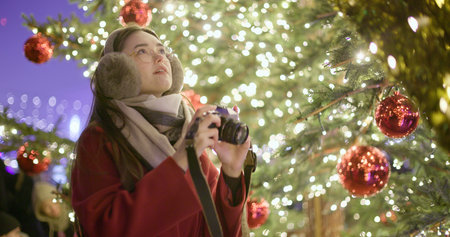 A young beautiful girl in a red coat stands near a Christmas tree with garlands and illuminations in the evening at the European Christmas Market and takes pictures with a vintage film camera.の写真素材