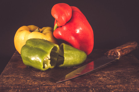 Green chopped pepper, with whole red and yellow peppers,  on a wooden chopping board with knife on black backgroundの写真素材