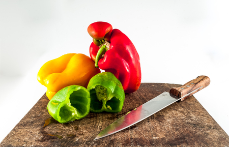 Green chopped pepper, with whole red and yellow peppers,  on a wooden chopping board with knife on white backgroundの写真素材