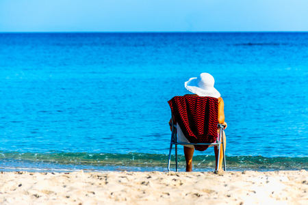 woman with white hat sitting on a chair on the beach in summer morningの写真素材