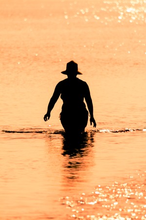 silhouette of woman with hat walking in the water of the sea at sunsetの写真素材