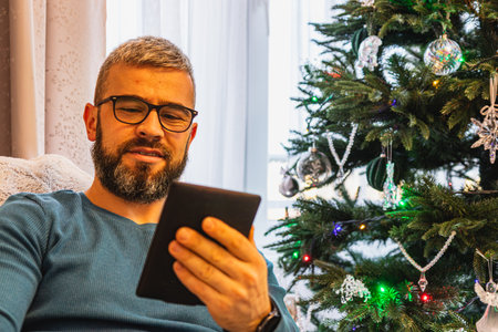 Enjoying the warmth of the holidays, a young man immerses himself in an e book while relaxing in a...