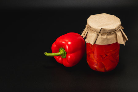 A jar of homemade pickled red pepper sits beside a fresh red pepper on a dark surface. This...