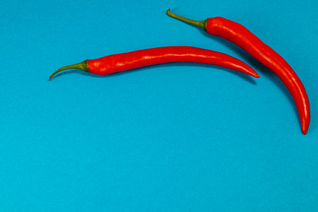 Two striking red chilies elegantly placed on a vivid blue background, showcasing their smooth...