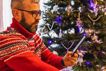 A young man enjoys reading an e book in a cozy chair, surrounded by the warm glow of Christmas lights on the tree. The festive atmosphere enhances the relaxed experience during the holiday season.の写真素材