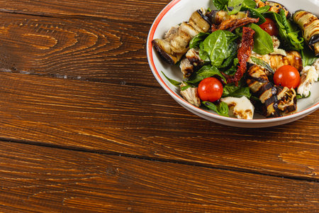Fresh salad features grilled eggplant, spinach, cherry tomatoes, and herbs, presented in a bowl. The setting includes a textured wooden tabletop, highlighting the meal's vibrant colors.の写真素材
