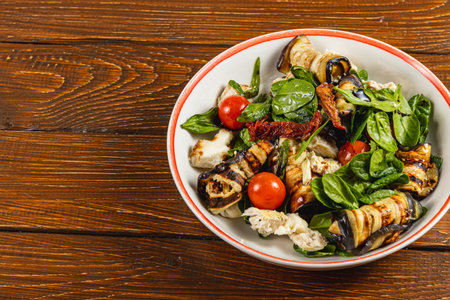 A vibrant bowl filled with a summer salad featuring grilled vegetables, cherry tomatoes, fresh spinach, and tender chicken pieces, all displayed on a rustic wooden table for a delightful meal.の写真素材