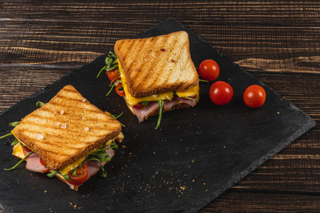 Two grilled sandwiches are served on a dark slate platter, filled with ham, double cheese, tomatoes, and greens, making a tempting choice for lunch or brunch. Fresh cherry tomatoes accompany the dish.の写真素材