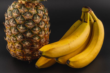 A vibrant arrangement of fresh pineapple and a bunch of ripe bananas sits on a dark surface. The natural textures and colors highlight the appeal of these fruits.の写真素材