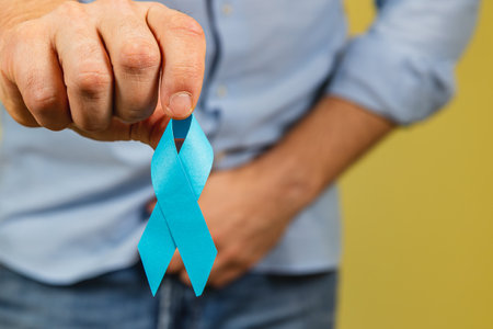 A man holds a blue awareness ribbon in front of his body against a yellow background, emphasizing the importance of cancer awareness and prevention in men's health.の写真素材