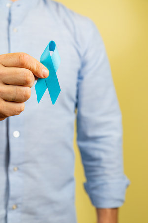 A person holds a blue ribbon in front of a yellow background, representing awareness for cancer...