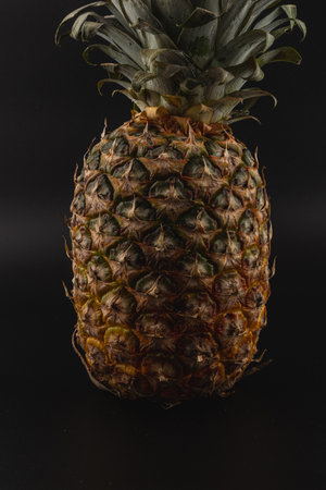 A ripe pineapple stands tall on a black surface. Its spiky, green leaves contrast with the rough,...