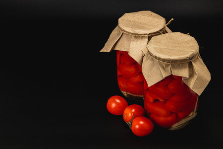 Two jars filled with vibrant pickled tomatoes sit on a dark surface alongside fresh, unprocessed cucumbers. This captures the essence of homemade preserving and freshness.の写真素材