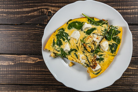 A delicious spinach and cheese frittata is served on a white plate, with a piece missing. The wooden...