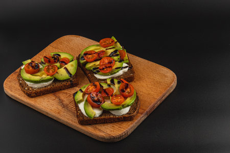 Three open-faced sandwiches made on dark bread are topped with slices of avocado, creamy cheese, and...