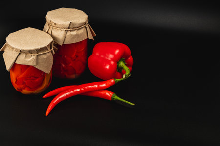 Jars of homemade canned food containing pickled tomatoes and sweet peppers sit on a dark surface. Fresh red peppers are placed beside the jars, showcasing vibrant colors.の写真素材