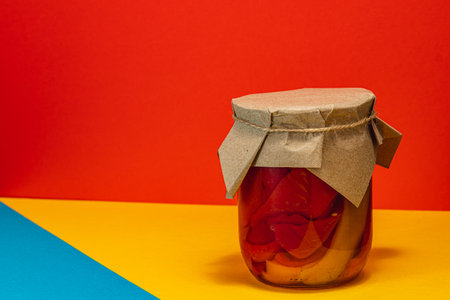 A jar filled with homemade pickled sweet peppers sits against a vibrant orange and yellow backdrop. The jar is sealed with paper, highlighting traditional canning practices.の写真素材