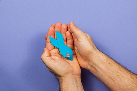 Two hands gently hold a blue ribbon, representing hope and support for cancer awareness. The background features a calming shade that emphasizes the ribbon's significance in promoting health.の写真素材