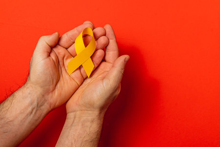 Two hands gently hold a yellow ribbon against a vibrant red background, representing hope and solidarity in the fight against cancer and the importance of awareness and support.の写真素材