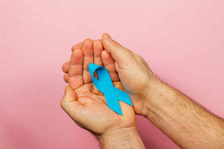 Male hands hold a blue ribbon against a pink backdrop, highlighting awareness for cancer prevention and encouraging support for patients undergoing treatment.の写真素材