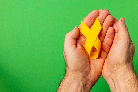 Two hands cup a yellow ribbon, a symbol of cancer awareness and support, against a bright green...