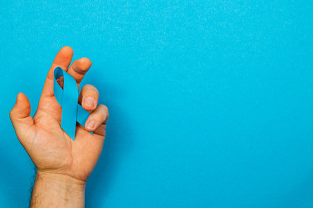 A male hand holds a blue ribbon against a bright blue background, symbolizing support for cancer awareness, diagnosis, and treatment. This represents courage and hope in the fight against illness.の写真素材