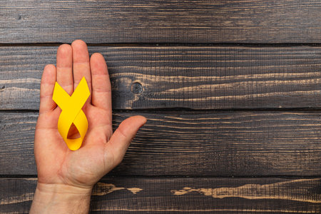 A person holds a yellow ribbon against a wooden background, symbolizing support for cancer awareness, courage, and solidarity in the fight against illness.の写真素材