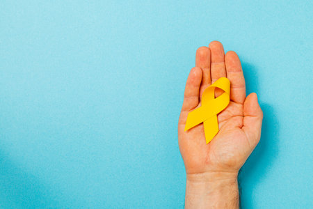 A hand holds a yellow ribbon against a blue background, representing hope and solidarity in the fight against cancer and a reminder of the importance of awareness and support.の写真素材