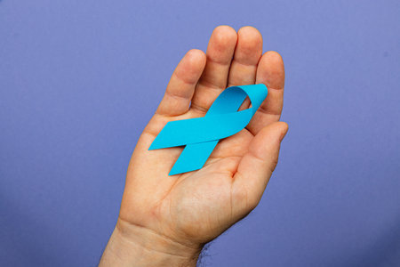 A male hand holds a symbolic blue ribbon against a purple background, promoting awareness for prostate cancer. This gesture emphasizes support, courage, and the importance of early detection.の写真素材