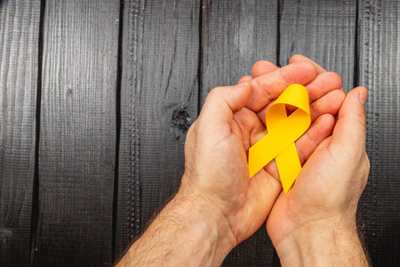 Two hands hold a yellow ribbon symbolizing hope and support in the fight against cancer. This act reflects solidarity and encouragement for those battling this illness.の写真素材