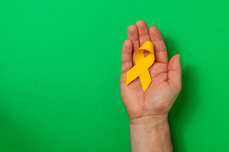A hand holds a yellow ribbon against a bright green background, representing awareness and solidarity in the battle against cancer and promoting mental health support.の写真素材