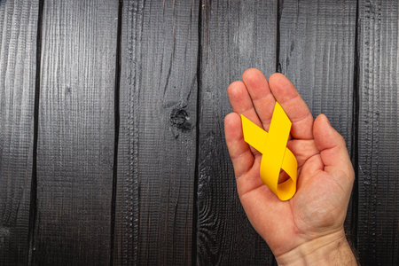 A female hand gently holds a yellow awareness ribbon, symbolizing hope and solidarity in the fight against cancer, set against a dark wooden background.の写真素材