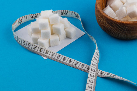 A pile of sugar cubes sits next to a wooden bowl on a blue surface. A measuring tape forms a heart...