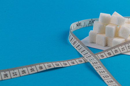 Sugar cubes are arranged neatly on a white plate, surrounded by a measuring tape. The bright blue background adds contrast, highlighting the focus on sugar and measurement.の写真素材