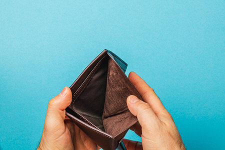 A man holds an empty wallet, reflecting despair and financial struggle against a blue background. His search for money highlights the challenges of poverty and budgeting in tough times.の写真素材