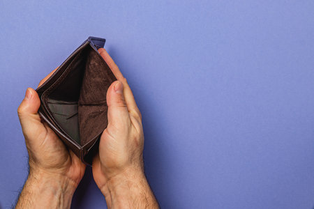 A male businessman holds an empty wallet in his hands, reflecting on his financial struggles. This...